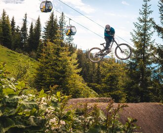 Mountain biker jumps high above forest ground at Bikepark Schladming, with cable cars in the background. | © Flowride