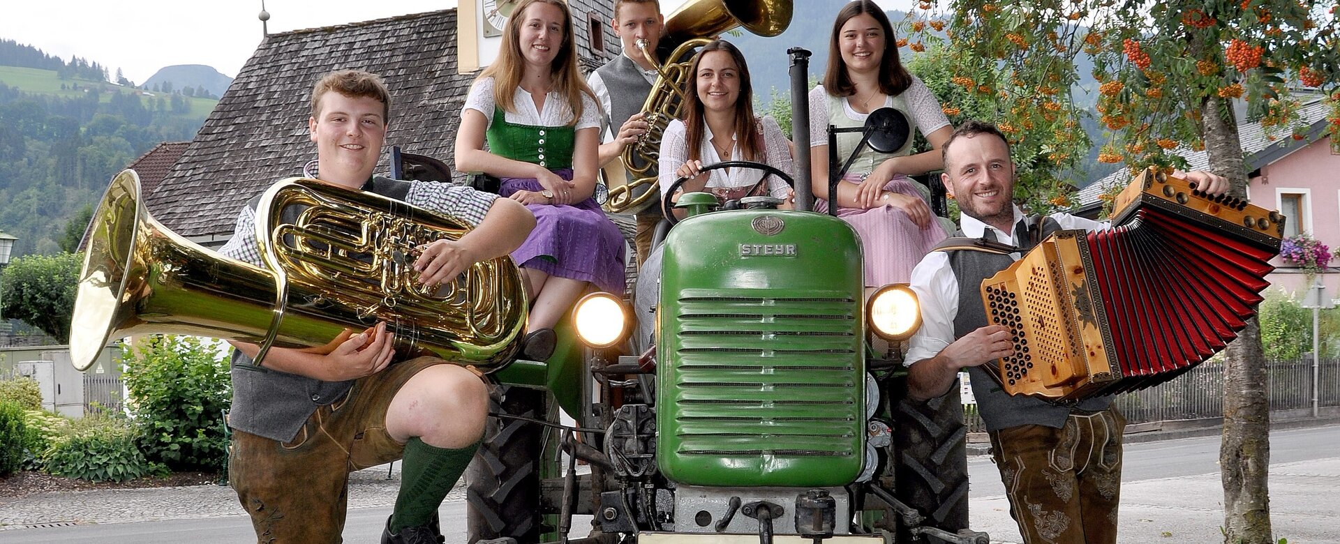 Gruppe junger Musikanten in Tracht mit Tuba, Harmonika & Co. auf grünem Steyr-Traktor beim Aicher Kirtag vor Kirchturm | © Aicher Kirtag