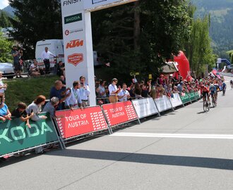 Cyclists cross the finish line at the Tour of Austria as spectators cheer by the roadside and finish arch | © JOSalzburg