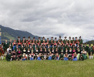 Large traditional group with two banners on alpine meadow, men in green, women in dirndl, children in front | © Trachtenverein