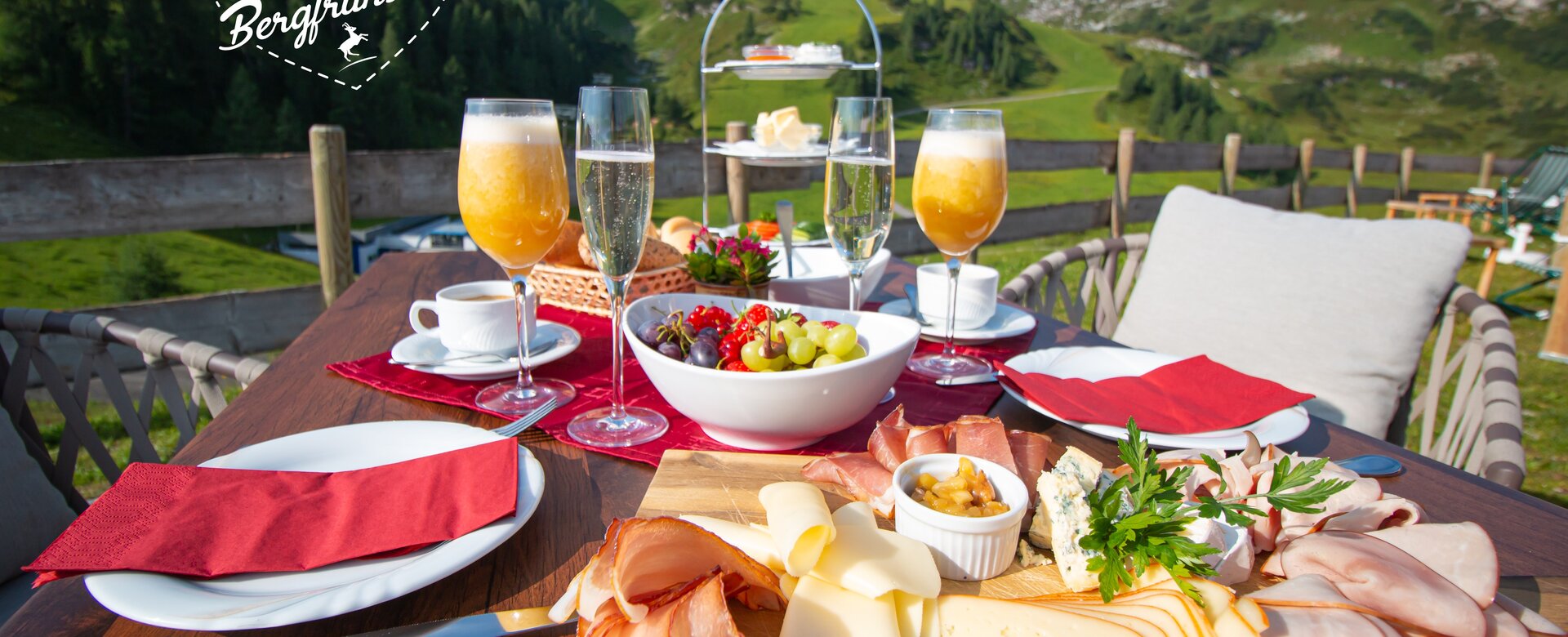Breakfast table with cheese, ham, fruit and drinks before sunny mountain scenery