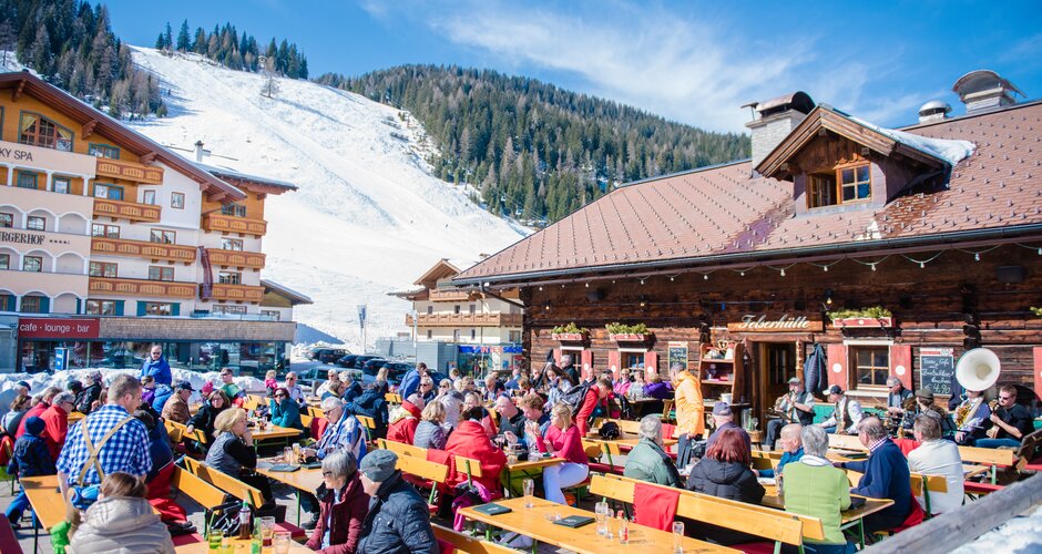 Ski terrace full of seated people and on the right you can see a wooden ski hut and behind the terrace a hotel and to the right of it the ski slope. | © Nadia Jabli Photography