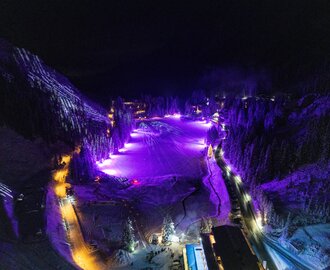 lluminated ski slope in mountain village at night with purple light show and snowy trees