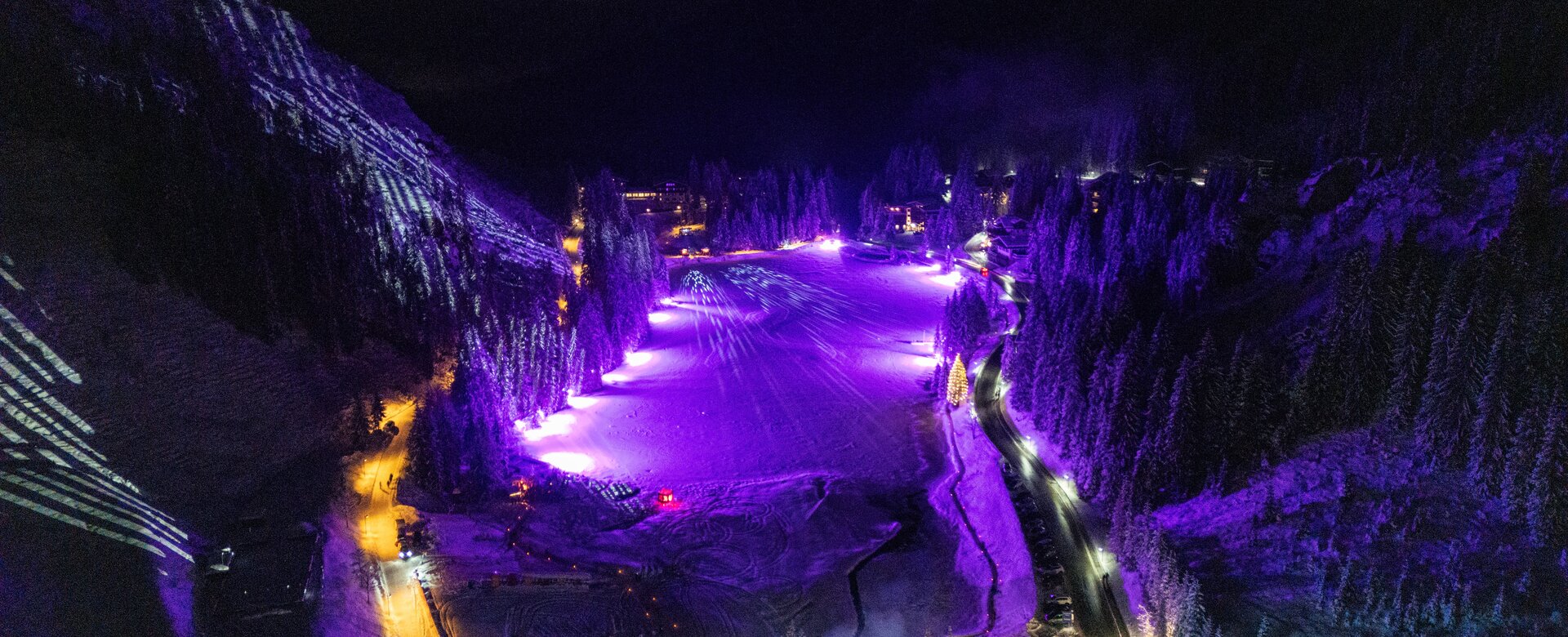 lluminated ski slope in mountain village at night with purple light show and snowy trees
