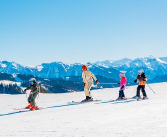 Ski instructor leads three children in colorful ski gear down a sunny slope. | © Hochkönig Tourismus GmbH