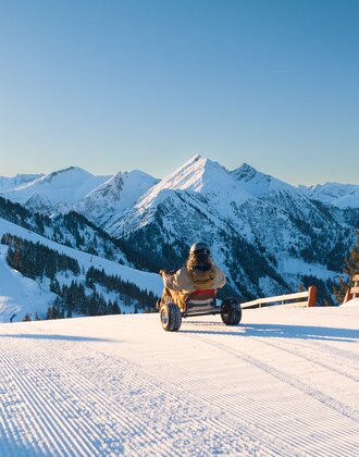Person rides snow kart on freshly groomed slope with view of snowy mountains in bright sunshine. | © Dorfgasteiner Bergbahnen AG