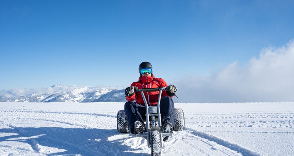 Person riding a mountaincart across snowy high plateau with alpine view and blue sky in Dorfgastein | © Dorfgasteiner Bergbahnen