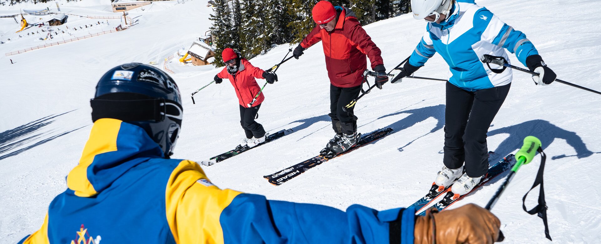 Ein Skilehrer in blau-gelben Skianzug zeigt drei Schülern eine Balanceübung mit ausgestreckten Armen.