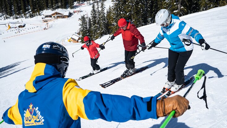 A ski instructor in a blue and yellow ski suit shows three students a balancing exercise with outstretched arms.