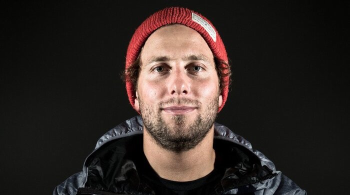 Josh Absenger looks at the camera wearing red beanie and dark jacket, captured against a solid black background.
