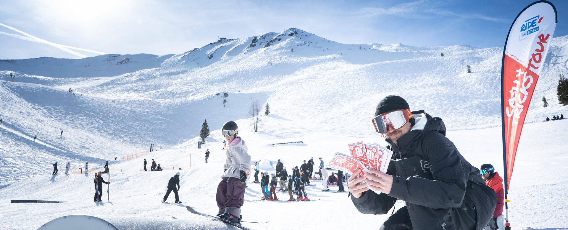 A judge kneels in snow holding red Cash4Tricks vouchers, spectators under bright alpine sun.