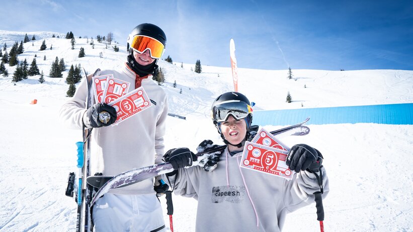 Two young freeskiers hold red Cash4Tricks vouchers, posing in snowpark scenery with skis and blue park tube behind them.