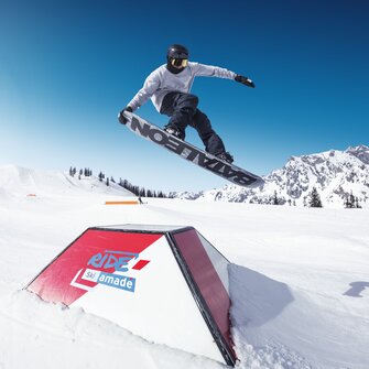 Snowboarder grabs board mid-jump over an obstacle in the RIDE Ski amadé snow park with blue sky and mountains.
