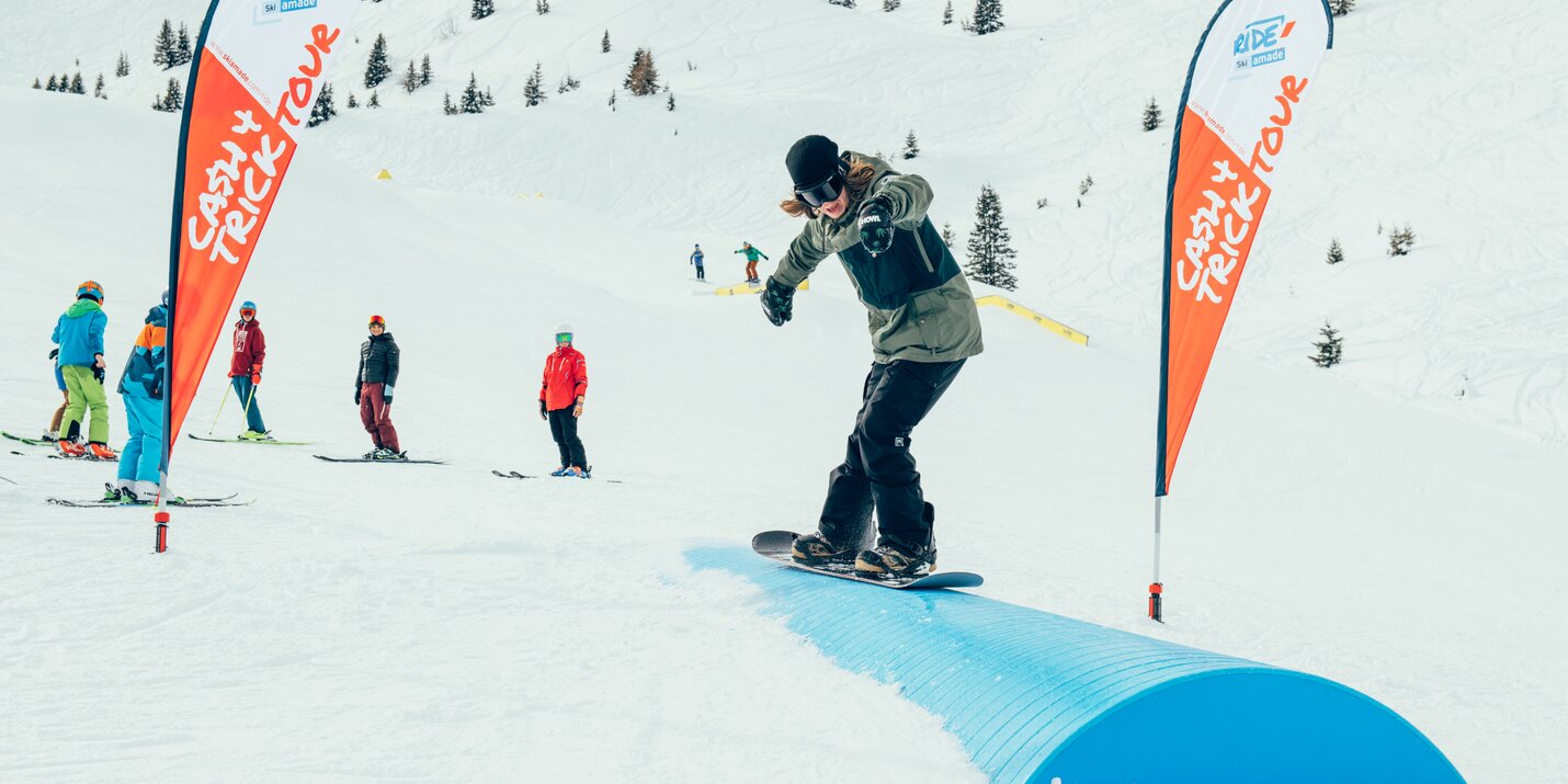 Snowboarder slides a blue tube at Cash4Tricks Gastein, with red flags and spectators standing on the snowy slope nearby.