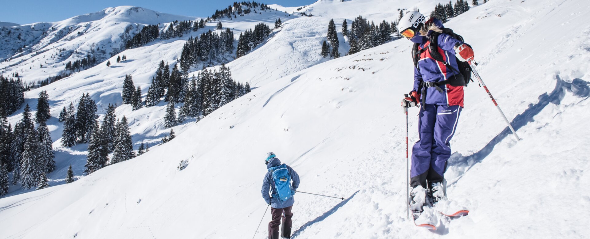 Two freeriders stand slightly offset and with their backs to each other on a slope and look down into the valley