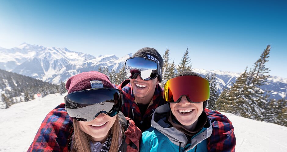 Three friends take a ski selfie with goggles on, smiling in front of snowy mountain scenery and bright blue sky.