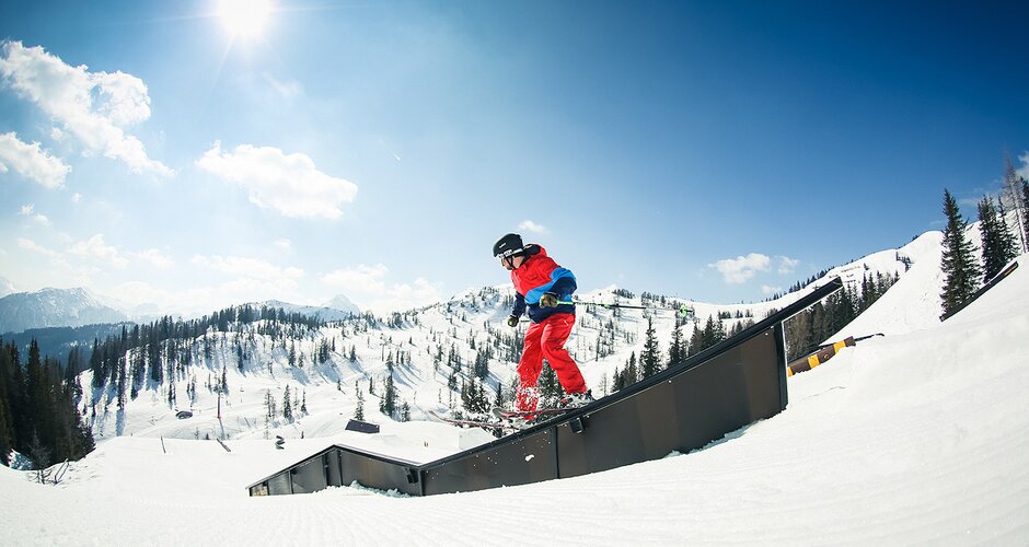 Freeskier in red-blue outfit slides on rail in snowpark with sunlight and snowy alpine scenery in background.