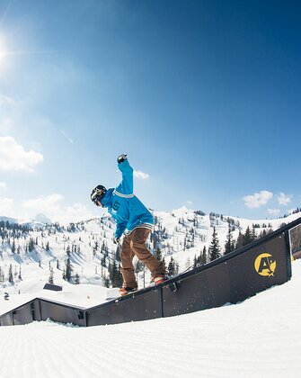 Snowboarder in blue hoodie slides on rail in sunny snowpark with snowy mountains and forest in the background.