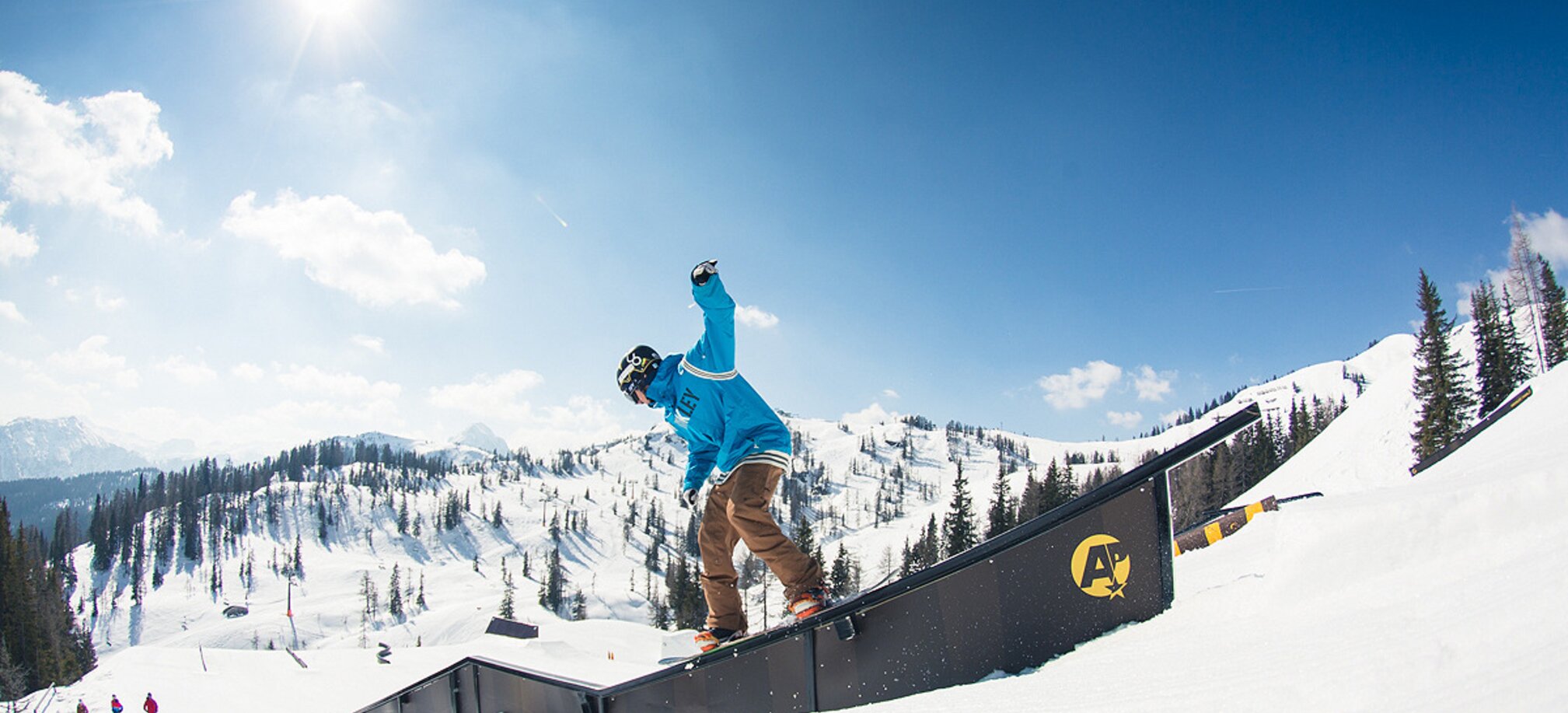 Snowboarder in blauem Hoodie slidet bei Sonne über Rail im Snowpark, verschneite Berge und Wald im Hintergrund.