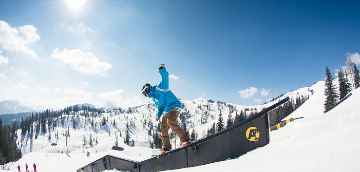 Snowboarder in blue hoodie slides on rail in sunny snowpark with snowy mountains and forest in the background.