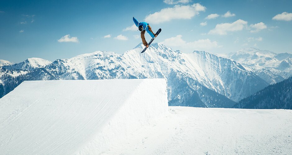Snowboarder in blue hoodie does grab trick above kicker in snowpark with snowy Alps and bright sunshine behind.