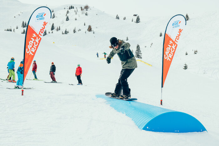 Snowboarder in green jacket balances on blue rail at Cash4Tricks Tour while skiers watch in the snowy background.