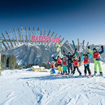 Group of kids on skis cheering under “Kidstag” arch in the Shuttleberg ski area on a sunny day | © Shuttleberg