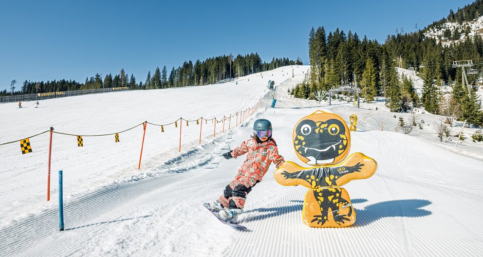 Kind mit Snowboard fährt bei Sonne auf der Funslope vorbei an bunter Figur im Skigebiet Shuttleberg | © Shuttleberg