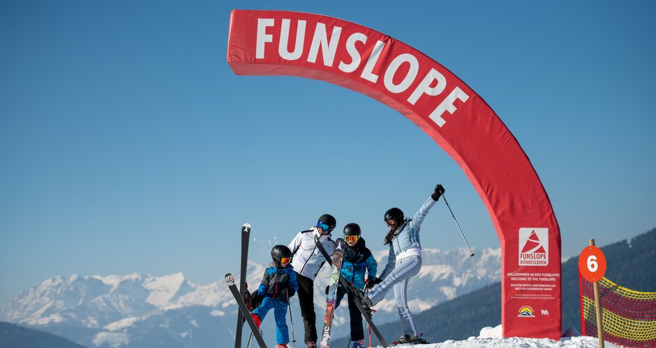 A family with two children stands under a red half arch on the Funslope and each holds a ski high in the air