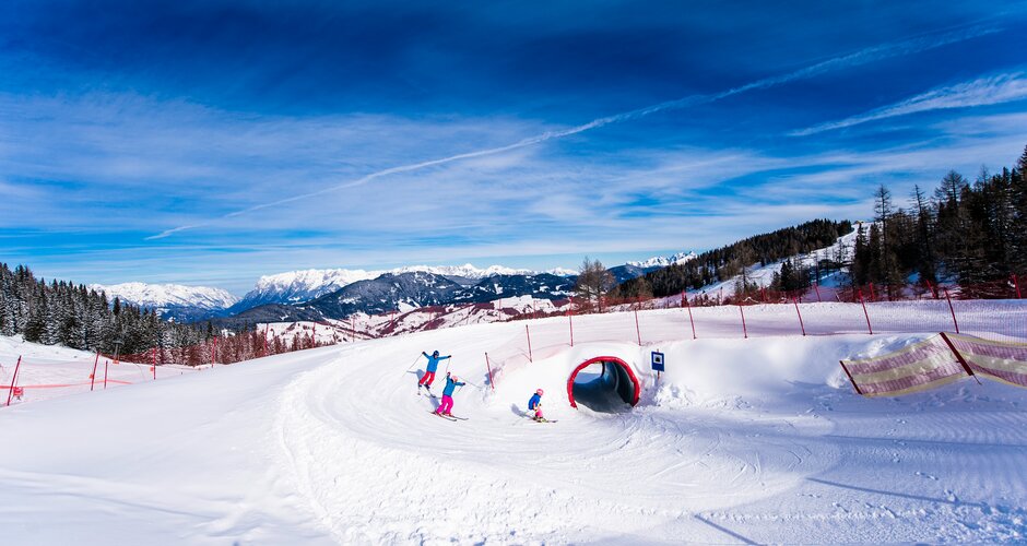 Three kids ski through a snow tunnel on the funslope, with sunshine and a mountain panorama in the background.