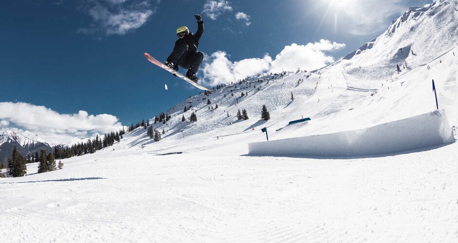 Snowboarder is in the middle of a jump and grabs the back edge of the snowboard with one arm and stretches the other high into the air | © Q-Parks Hannes Mautner