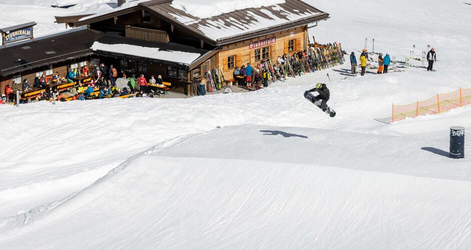 Snowboarder jumps over a ski jump and in the background spectators stand in front of a ski hut | © Q-Parks Hannes Mautner