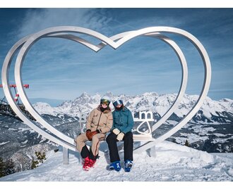 Two skiers sit in the Styrian Heart frame on Reiteralm with Dachstein mountains behind