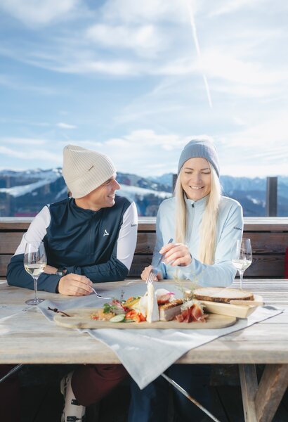 Two people eat snack with cheese, ham, bread and wine on ski hut terrace with mountain backdrop | © Ski amadé