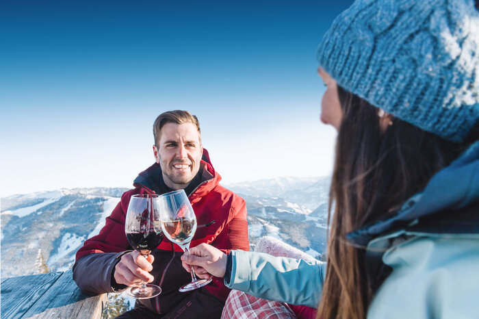 A man in a red ski jacket smiles and clinks glasses with a woman in a blue cap and blue ski jacket as they sit at a wooden table