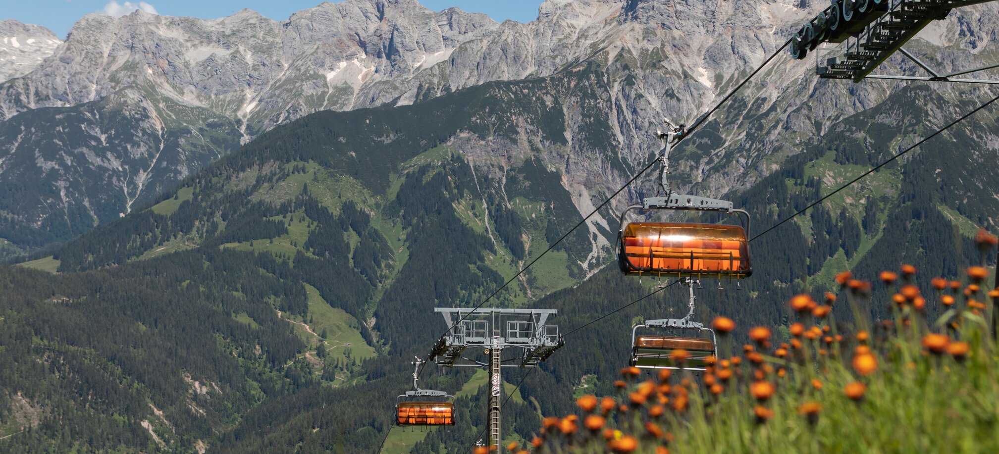 Orange chairlifts of Hochmaisbahn float above alpine slopes and blooming meadows in summer near Hochkönig. | © Roland Haschka