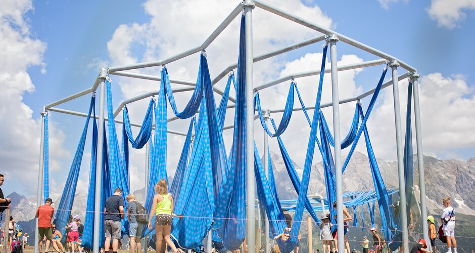 Metal frame with blue fabric loops that can be used as a swing and children playing with it | © Manu Lochner