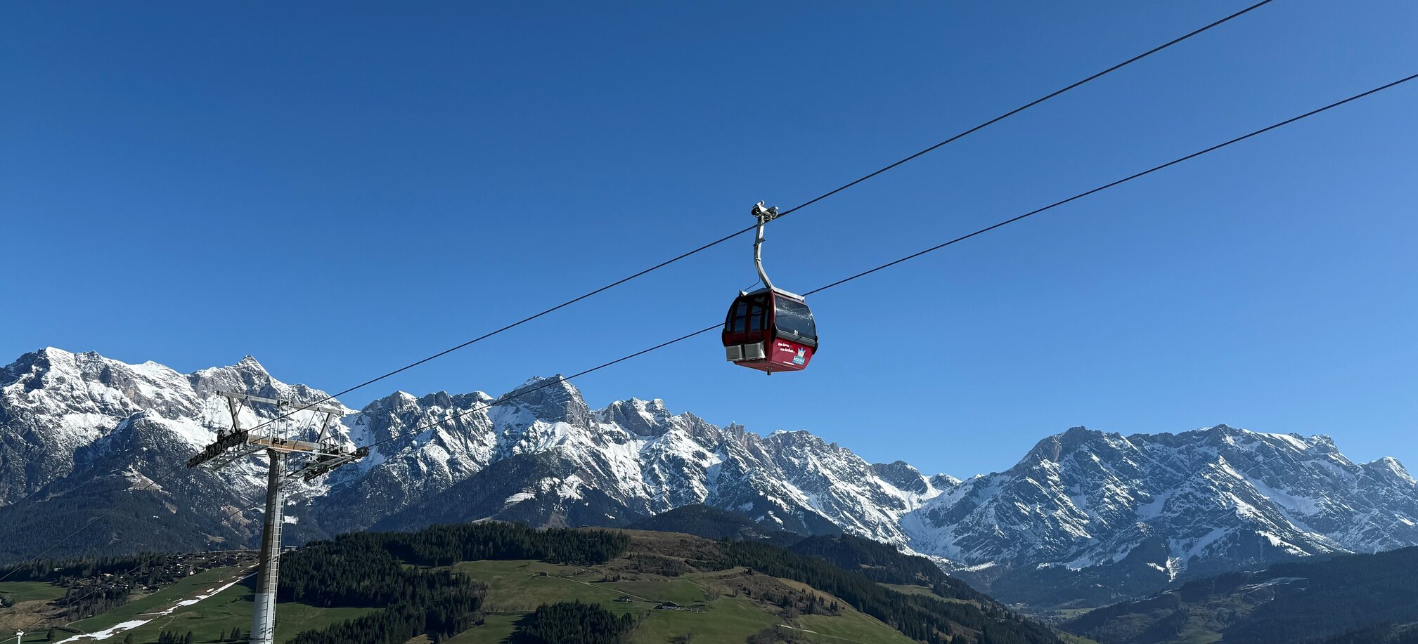 Red Abergbahn gondola glides in summer above green hills and snow-covered Hochkönig range under a clear blue sky. | © Aberg - Hinterthal - Bergbahnen AG