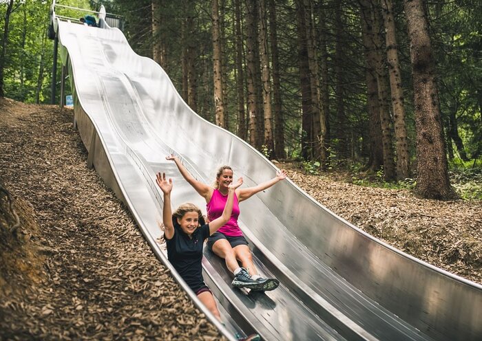 Girl and woman laughing with raised arms while sliding down the twin slide at the forest slide park Natrun.
