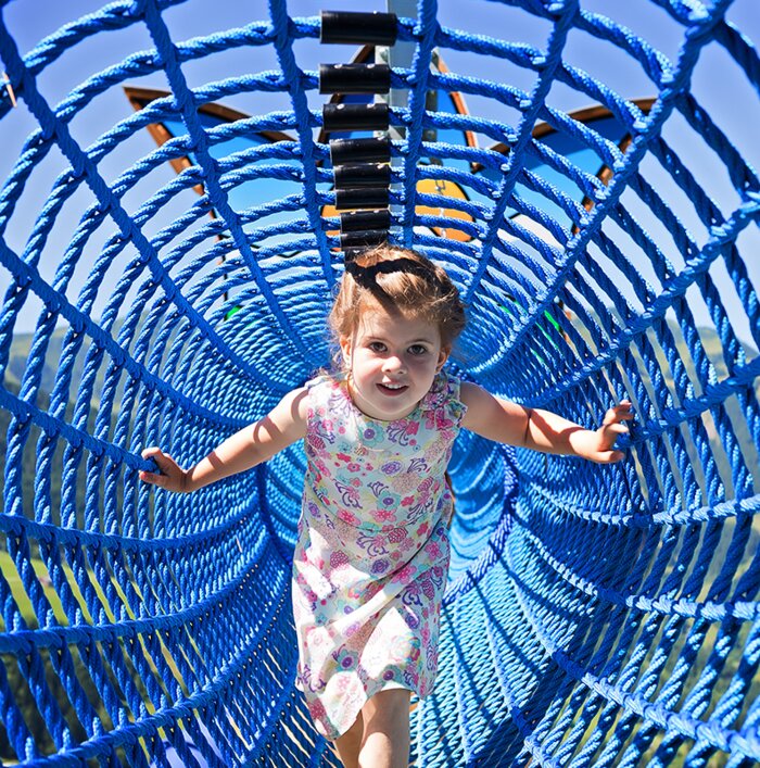 Little girl in a summer dress climbs through a blue rope tunnel on an adventure playground with scenic mountain views. | © Hochkönig Tourismus GmbH