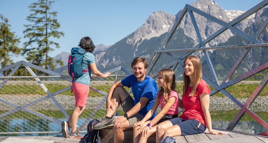 Four hikers take a break on a platform at Lake Prinzensee in Maria Alm with a scenic view of the Hochkönig mountains. | © Roland Haschka