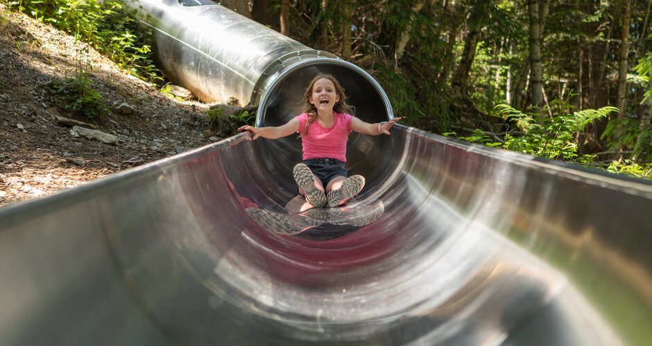 Laughing girl slides down a long stainless steel slide at the shady forest slide park on Prinzenberg Natrun. | © Roland Haschka
