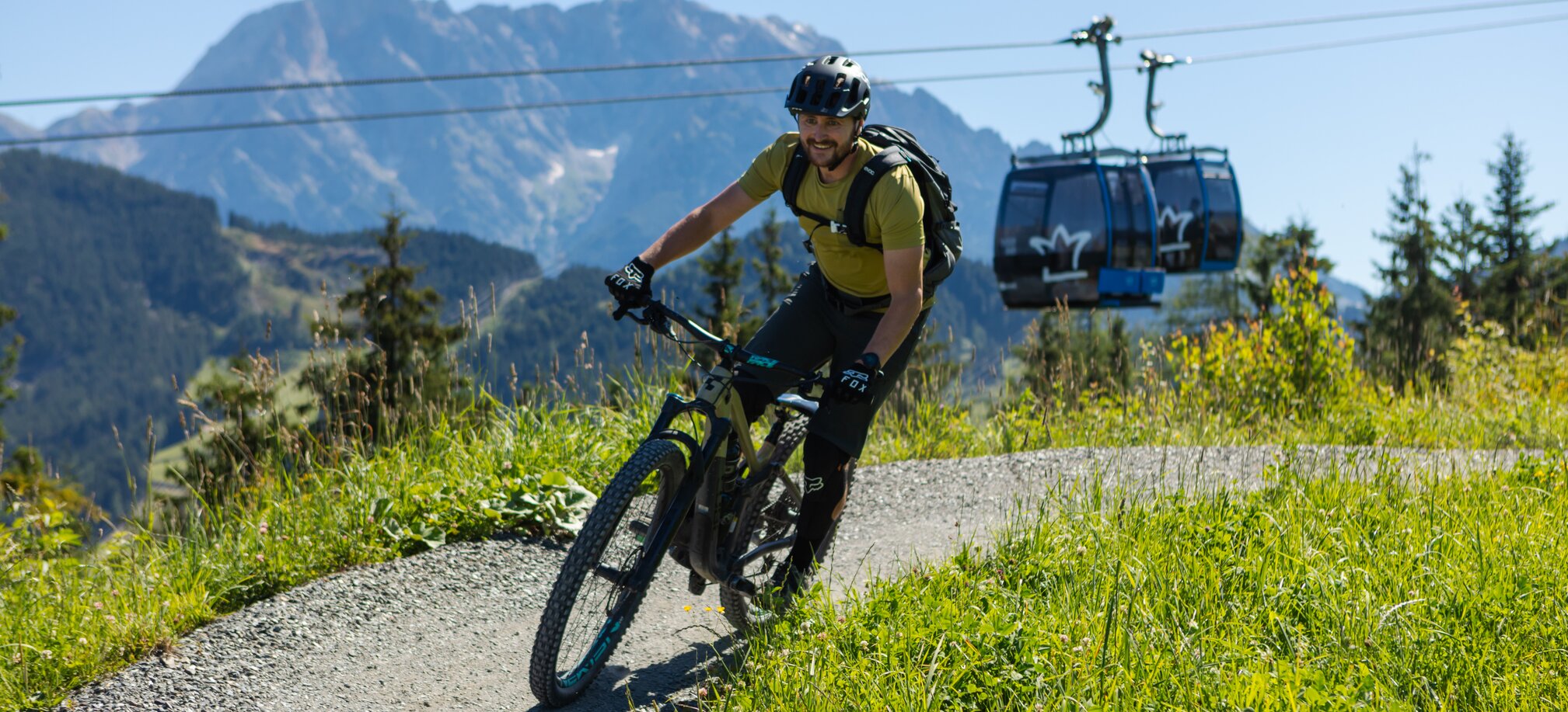 A mountain biker rides a winding trail with a gondola lift and scenic alpine peaks in the background under a clear blue sky. | © Roland Haschka
