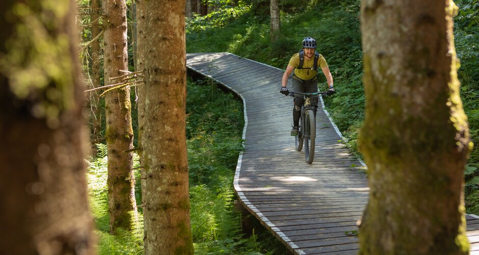 Mountain biker with helmet and gear rides a winding wooden trail through dense forest with sunlight filtering through trees. | © Roland Haschka