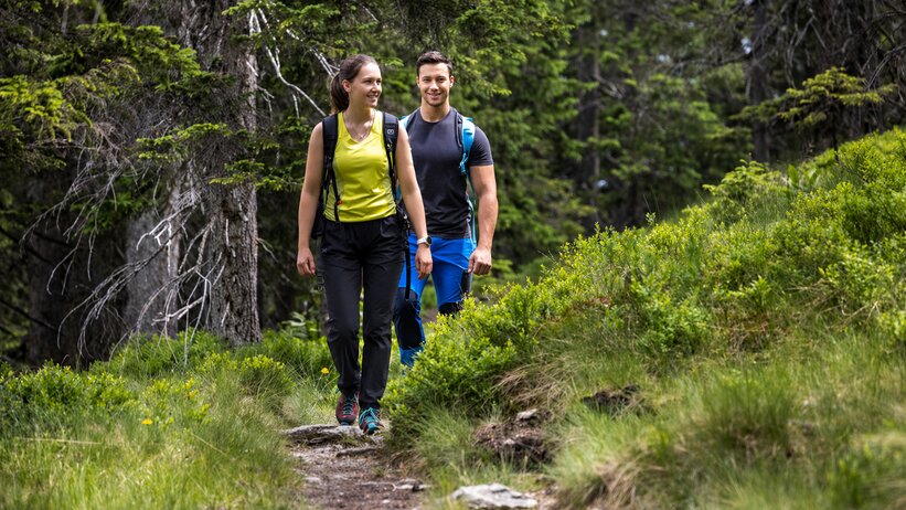 Two hikers walk along a narrow forest trail through green meadows and dense conifer trees in Filzmoos. | © filzmoos.ski_Christian Fischbacher