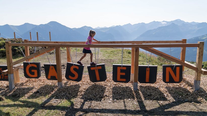 Girl balancing on playground blocks spelling GASTEIN with Alpine backdrop in Gastein Valley | © Gasteiner Bergbahnen AG