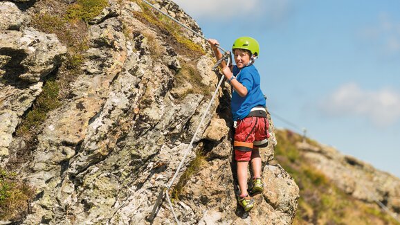 Boy with helmet and harness climbing on secured rock face in sunny alpine landscape | © Gasteiner Bergbahnen AG