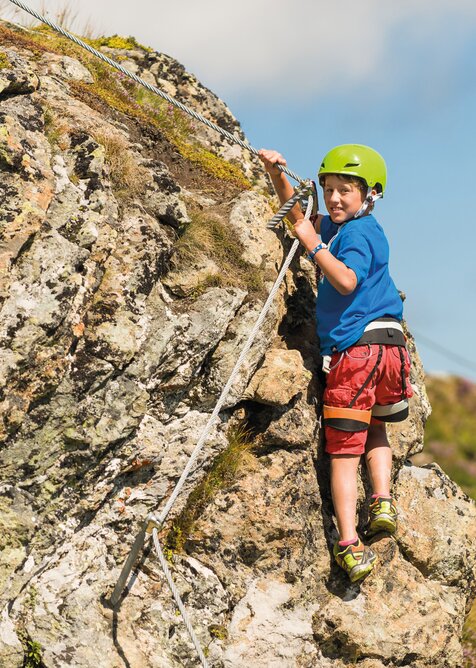 Boy with helmet and harness climbing on secured rock face in sunny alpine landscape | © Gasteiner Bergbahnen AG