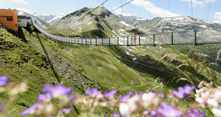 Two people on Stubnerkogel suspension bridge with blooming purple summer flowers in the foreground | © Gasteiner Bergbahnen, Marktl Photography