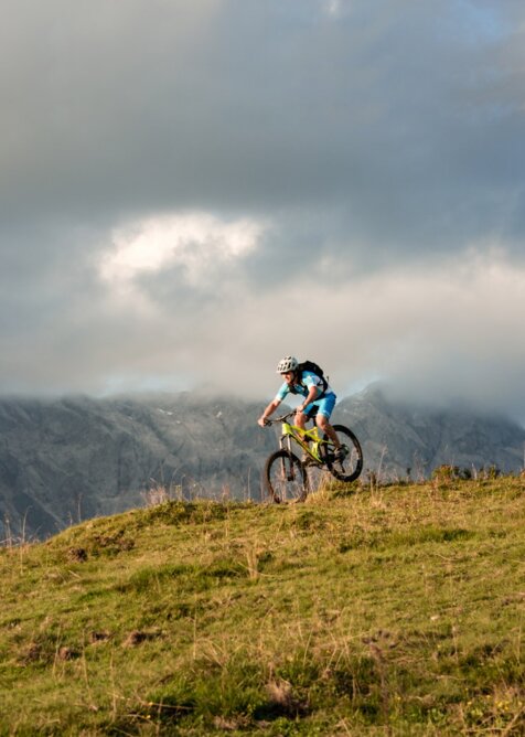 Mountain biker rides alpine meadow with cloud-covered Hochkönig range in evening light | © Christian Schartner